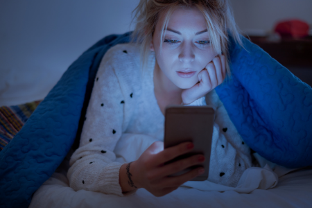Young woman looking at the blue light from her iphone screen, preventing her from getting better sleep.
