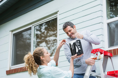 A couple with a can of paint working on the exterior of their home.