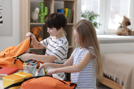 Two kids packing their backpacks at a table designated as part of the back-to-school home organization plan.